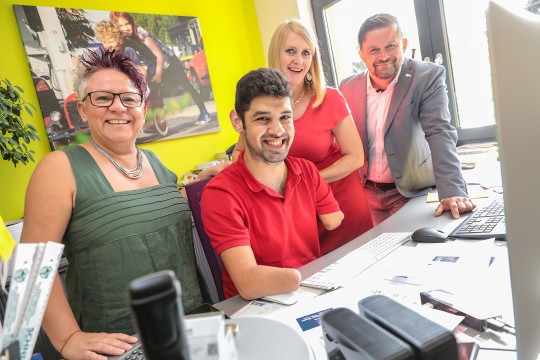 Auf dem Bild sieht man eine Beratungssituation am Arbeitsplatz. Alle lächeln in die Kamera.